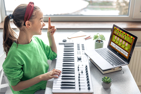 Photo of a little girl with an electronic keyboard, learning piano through an app on a laptop. The screen on the app displays an illustration of a carnival-style shooting gallery, with a row of ducks and a bar of sheet music.