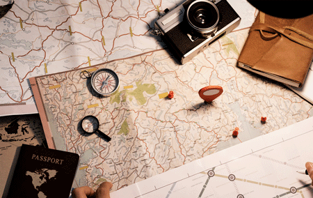 Photo of classic travel images on a desk or table: maps, passport, compass, magnifying class, travel diary, and a camera.